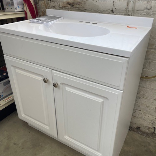 Close-up of our display of vanities, featuring a white vanity with a white sink and countertop, white cabinets with silver hardware, and a drawer.